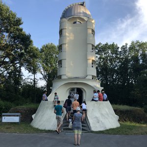 Frontalansicht. Menschen, die an einem sonnigen Tag im Eingangsbereich des Einsteinturms auf einer Mauer sitzen oder gerade hineingehen.