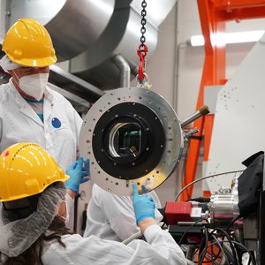 Two persons with helmet, face masks and white coats work on a round object with a lens in the middle, which is hold by a crane.