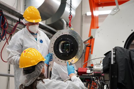 Two persons with helmet, face masks and white coats work on a round object with a lens in the middle, which is hold by a crane.