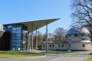 The Babelsberg research campus.On the left, the Schwarzschildhaus with its glass façade and peaked roof; on the right, the historic metal-domed building of the library.  In the background, the white main dome of the Humboldthaus is visible through trees.