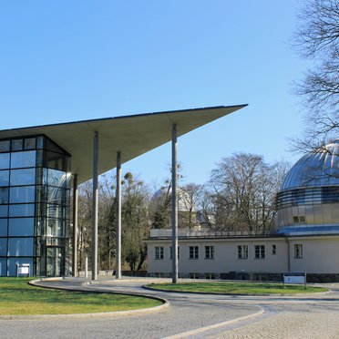 The Babelsberg research campus.On the left, the Schwarzschildhaus with its glass façade and peaked roof; on the right, the historic metal-domed building of the library.  In the background, the white main dome of the Humboldthaus is visible through trees.