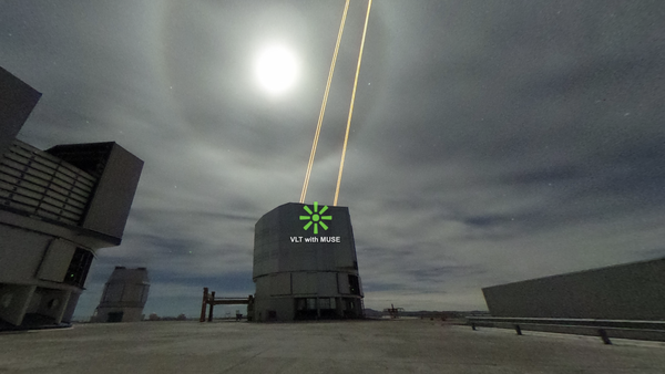 Screenshot der VR-Umgebung. Zu sehen ist das kastenförmige Very Large Teleskop neben einem weiteren Teleskop vor einem bewölkten Himmel. Oben schießen zwei Laserstrahlen heraus.