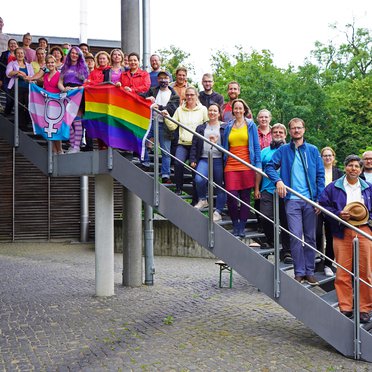 Various AIP staff members in colourful clothing stand on a staircase with a rainbow flag and a transgender flag.