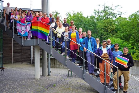 Various AIP staff members in colourful clothing stand on a staircase with a rainbow flag and a transgender flag.