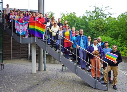 Various AIP staff members in colourful clothing stand on a staircase with a rainbow flag and a transgender flag.