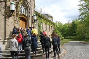 A group of schoolgirls, visible from behind, walk through the large wooden door of the Humboldthaus. One of its two white side domes is visible. The house itself is overgrown with ivy.