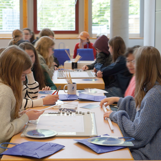 Girls sitting at a table with crating materials, among them a white board, folding rule, wooden skewers, and star charts