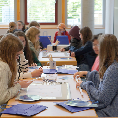 Girls sitting at a table with crating materials, among them a white board, folding rule, wooden skewers, and star charts