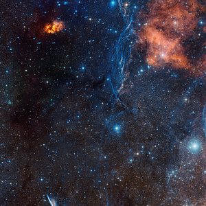 Sternenhimmel mit zahlreichen Sternen, orangefarbenen Nebelwolken und bläulichen Gasfilamenten im Weltraum.