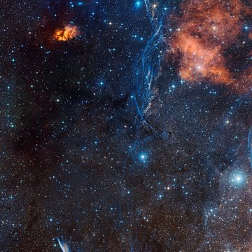 Sternenhimmel mit zahlreichen Sternen, orangefarbenen Nebelwolken und bläulichen Gasfilamenten im Weltraum.