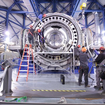 Persons with safety helmets work on the backside of a telescope in a big hall