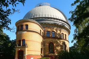 Der Große Refraktor von außen an einem sonnigen Tag. Seitliche Ansicht. Die weiße Kuppel ist einen Spalt geöffnet, die Bäume werfen Schatten auf den Klinkerbau.