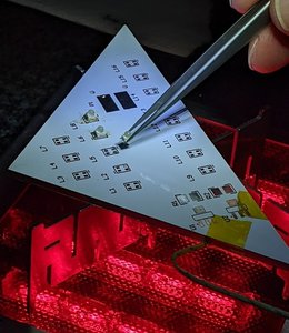 Tweezers place a tiny light-emitting diode at a marked point on the workpiece, which is held above red glowing heating elements